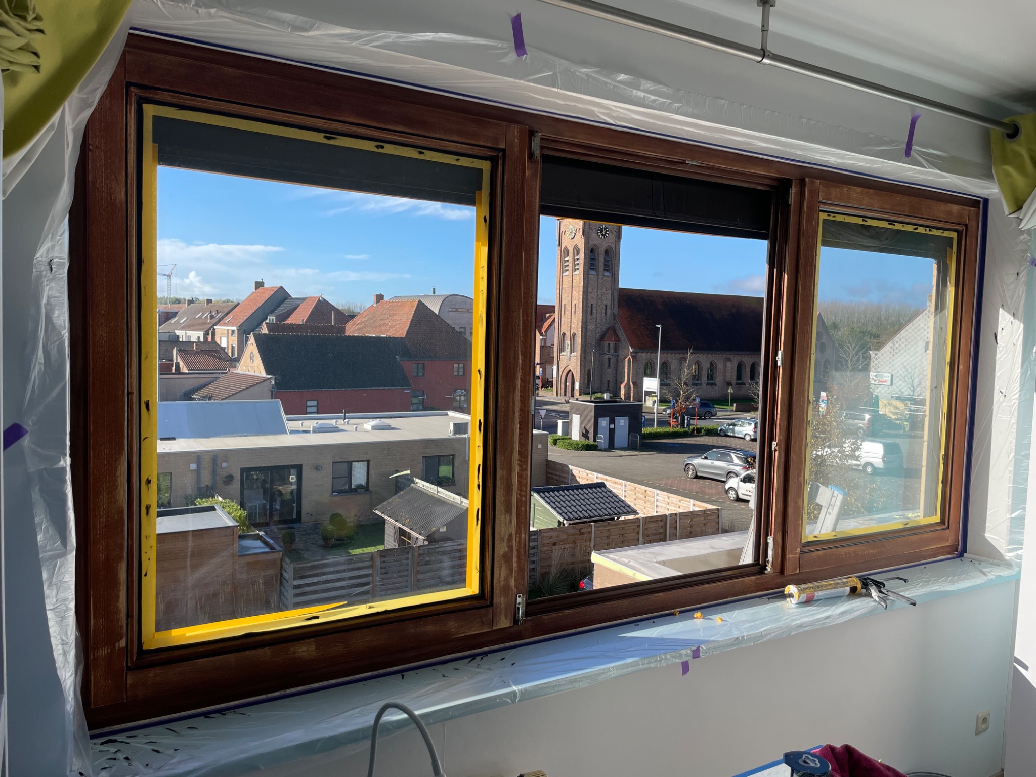 Drewniane okno z żółtą taśmą malarską zabezpieczającą szyby podczas renowacji, widok na budynki mieszkalne i kościół w tle.
