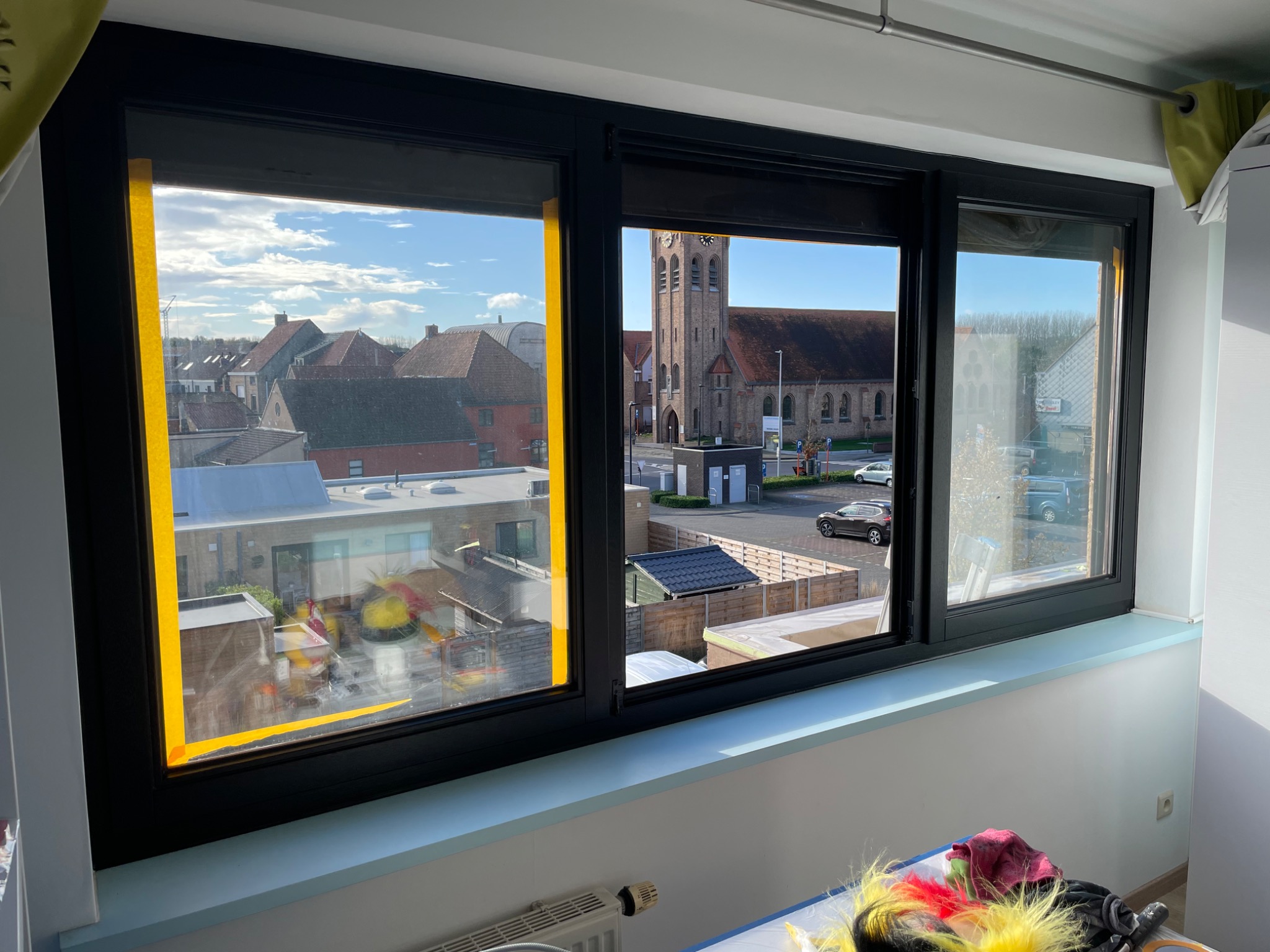 Nowe, czarne okno trzyskrzydłowe z widokiem na kościół i zabudowania mieszkalne, zabezpieczone żółtą taśmą malarską podczas montażu.