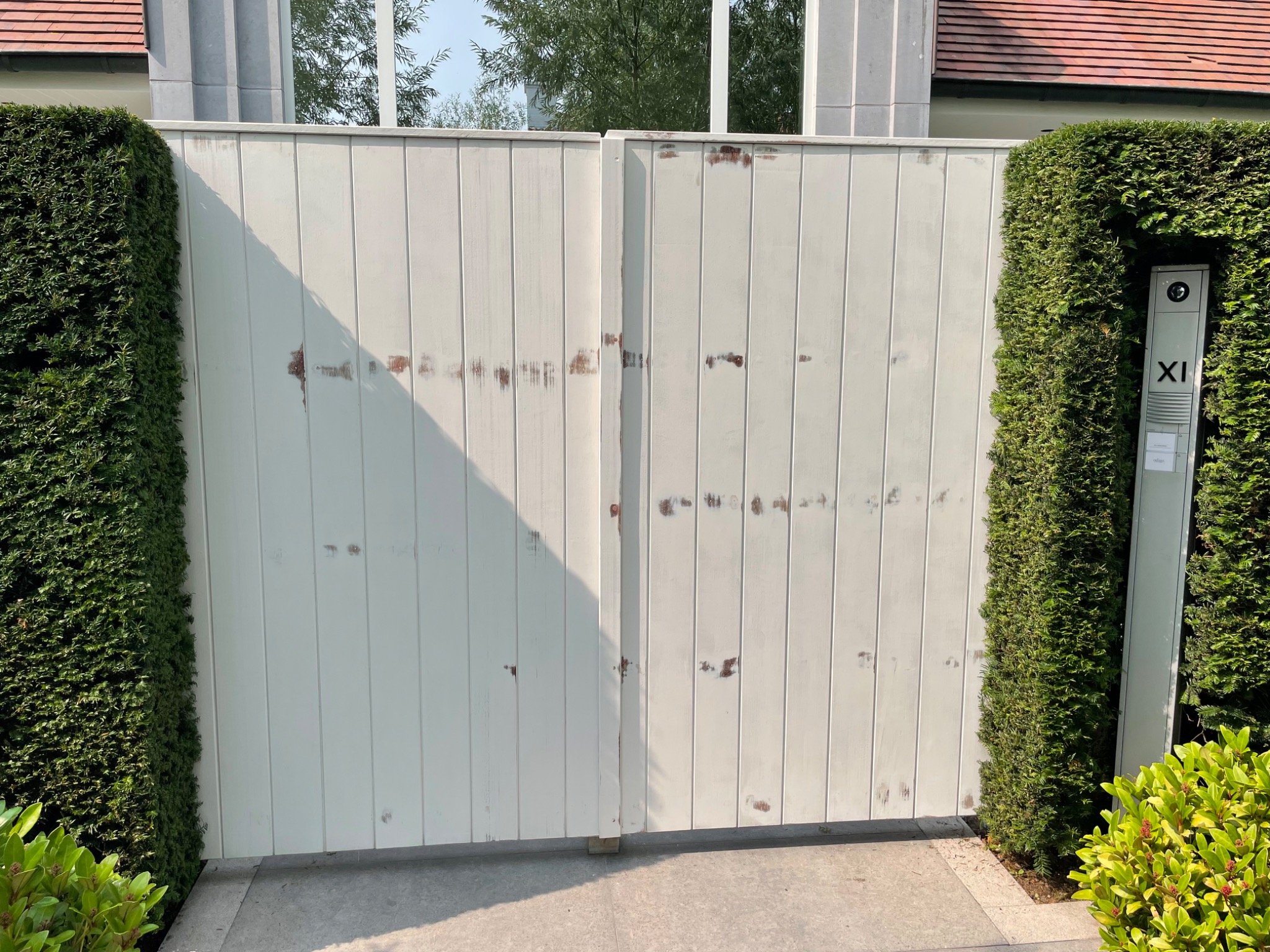 Biała, drewniana brama wjazdowa z widocznymi śladami użytkowania i odpryskami farby, otoczona zielonymi krzewami i z numerem domu 'XI' na słupku.