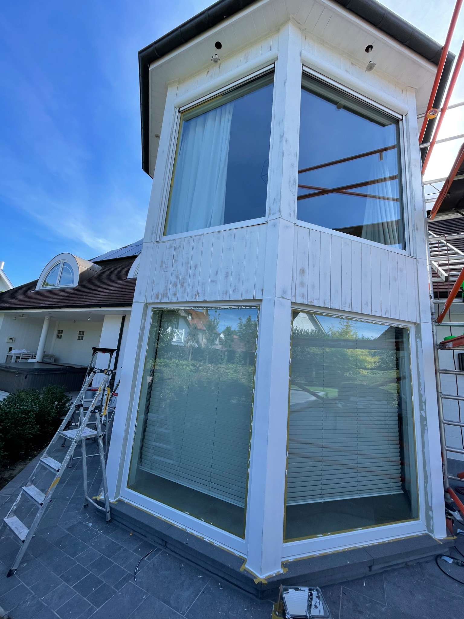 Wysoka, przeszklona weranda z dużymi oknami, częściowo pomalowana na biało, z widocznymi śladami wcześniejszych warstw farby, drabina oparta o ścianę, w tle dom z oknem w kształcie półkola.