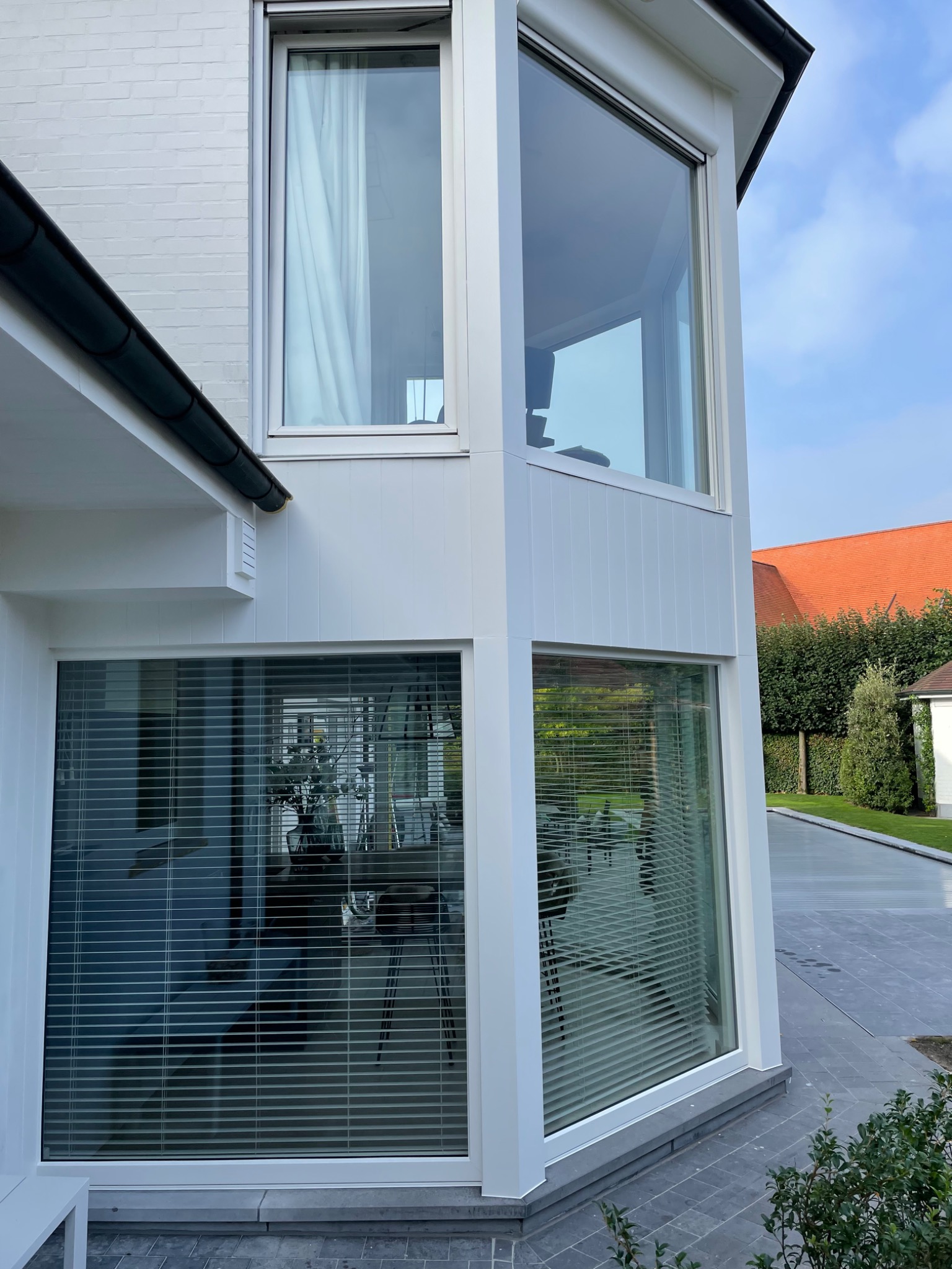 Nowoczesny narożnik domu z dużymi oknami z białymi ramami i żaluzjami, widok z zewnątrz na elewację i otoczenie.