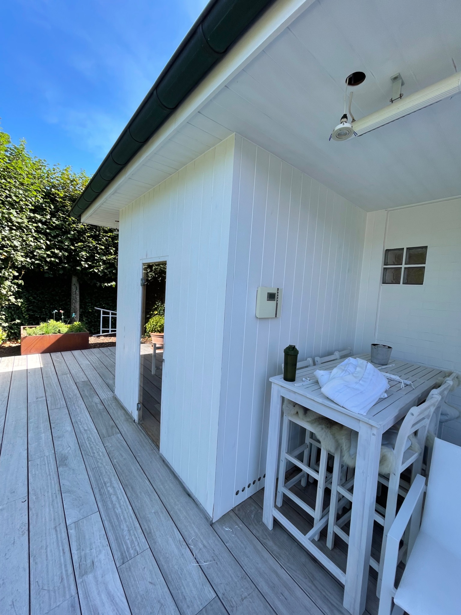 Biała altana z drewnianą podłogą i stołem z krzesłami, widok na zewnątrz przez szklane drzwi, oświetlenie sufitowe