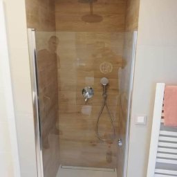 Perfect Styl Sp. Z o.o - Kabina prysznicowa z drewnopodobnymi płytkami, deszczownicą i odpływem liniowym, widoczna przez szklane drzwi, obok grzejnik drabinkowy z różowym ręcznikiem.