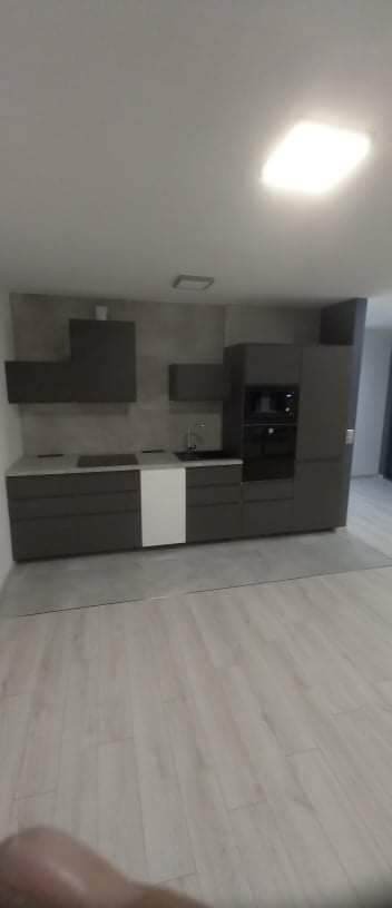 Nowoczesna kuchnia w odcieniach szarości z białym akcentem na jednej z szafek, oświetlona górnym światłem, z drewnopodobną podłogą panelową i szarą ścianą nad blatem.