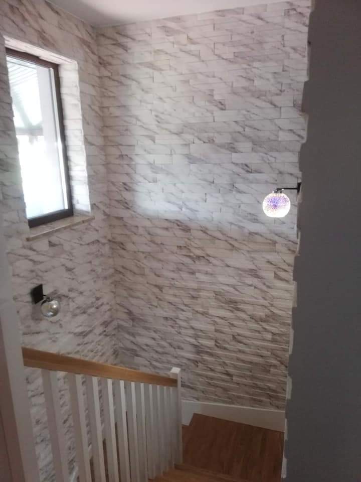 Klatka schodowa z drewnianymi schodami i białą balustradą, ściany wyłożone panelami imitującymi biały marmur, oświetlenie w postaci okrągłych kinkietów.
