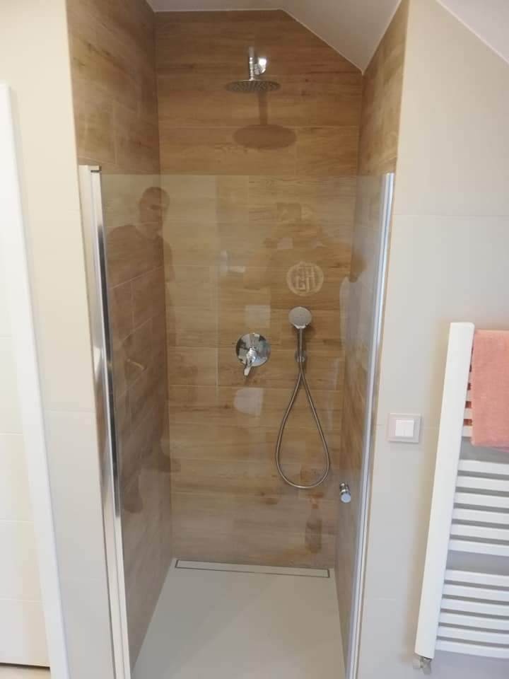 Kabina prysznicowa z drewnopodobnymi płytkami, deszczownicą i odpływem liniowym, widoczna przez szklane drzwi, obok grzejnik drabinkowy z różowym ręcznikiem.