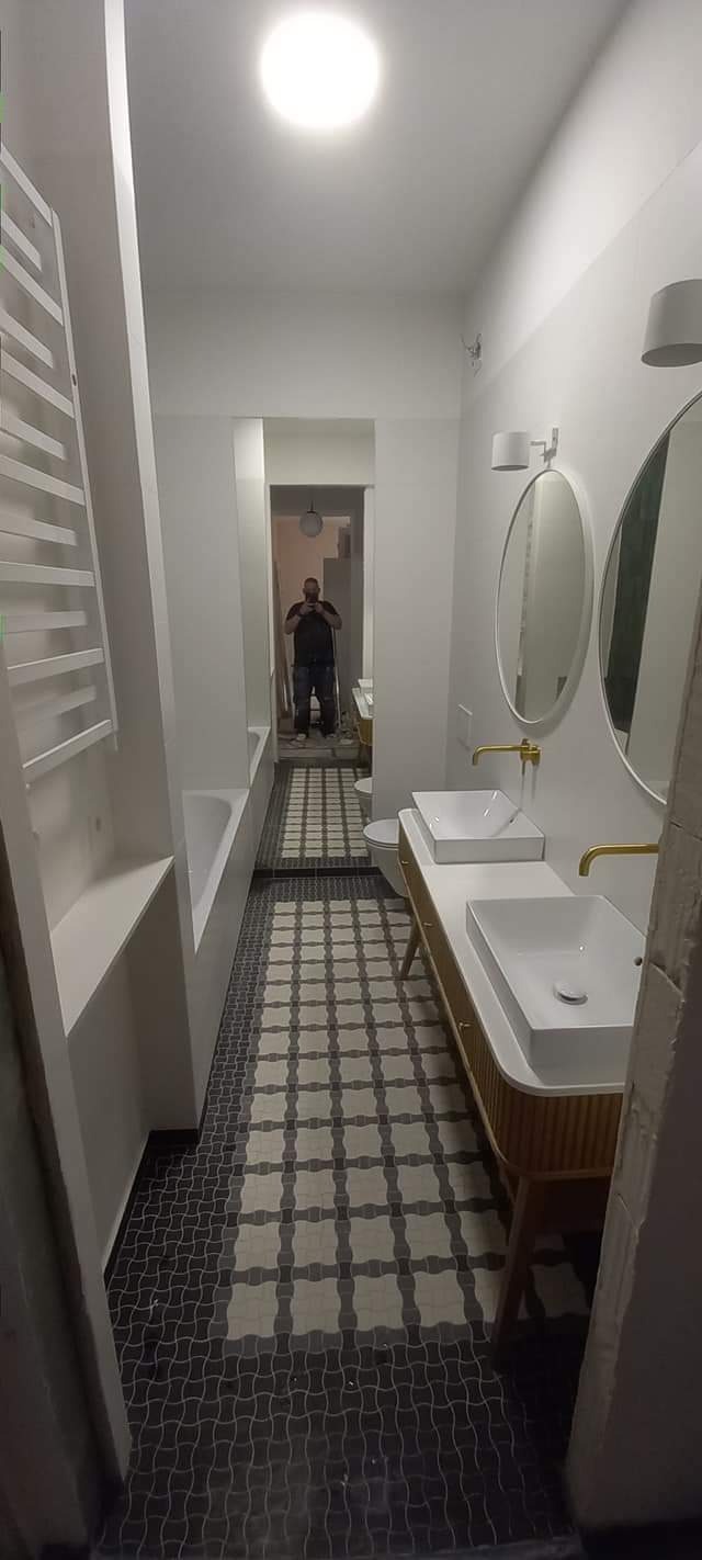 Wąska łazienka w Warszawie po remoncie, z dwoma umywalkami na drewnianej szafce, owalnymi lustrami, grzejnikiem drabinkowym i wzorzystą podłogą z płytek, widok z perspektywy wejścia, w głębi...