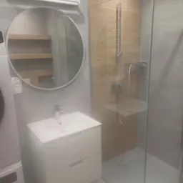 Nowoczesna łazienka z kabiną prysznicową typu walk-in, okrągłym lustrem z podświetleniem i białą szafką umywalkową z dwiema szufladami, widoczna pralka obok umywalki.