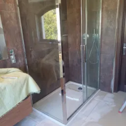 Kabina prysznicowa z przesuwnymi drzwiami, chromowanymi profilami i deszczownicą, zamontowana w łazience z brązowymi płytkami ściennymi. Na podłodze widoczne ślady po fugowaniu, a obok kabiny stoi...