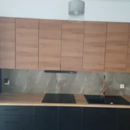 Nowoczesna kuchnia z drewnianymi górnymi szafkami, czarnymi dolnymi szafkami, marmurowymi płytkami na ścianie i miedzianą baterią.