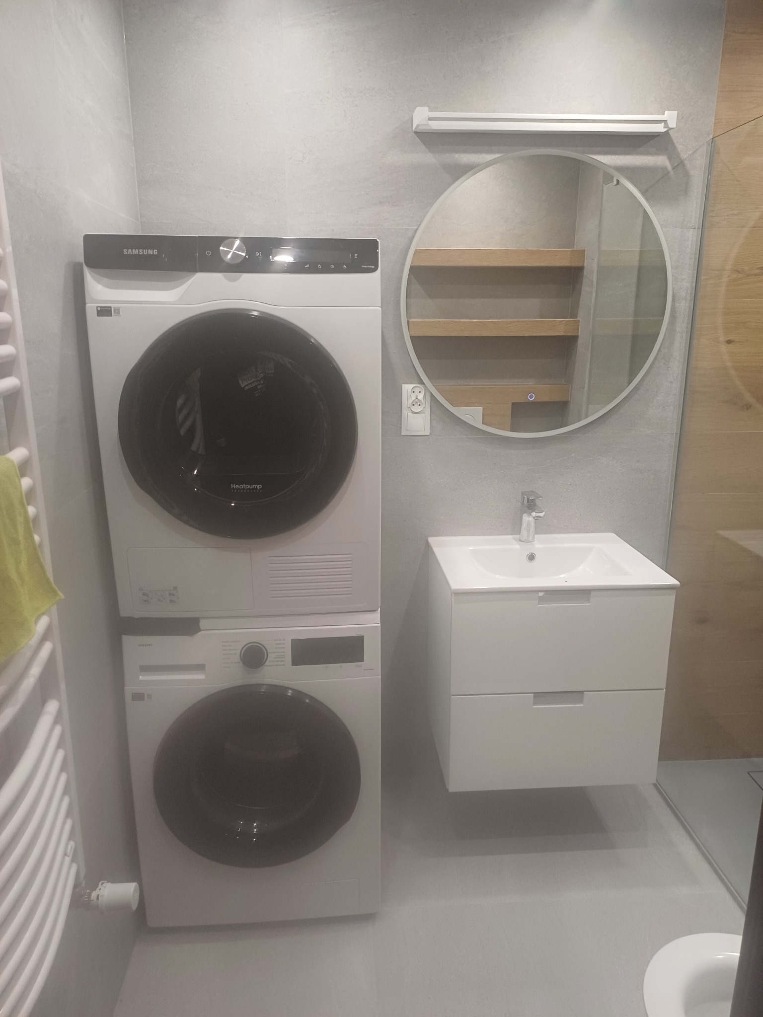 Nowoczesna łazienka w szarościach z pralką i suszarką Samsung ustawionymi jedna na drugiej, okrągłym lustrem z podświetleniem LED i białą szafką pod umywalką.