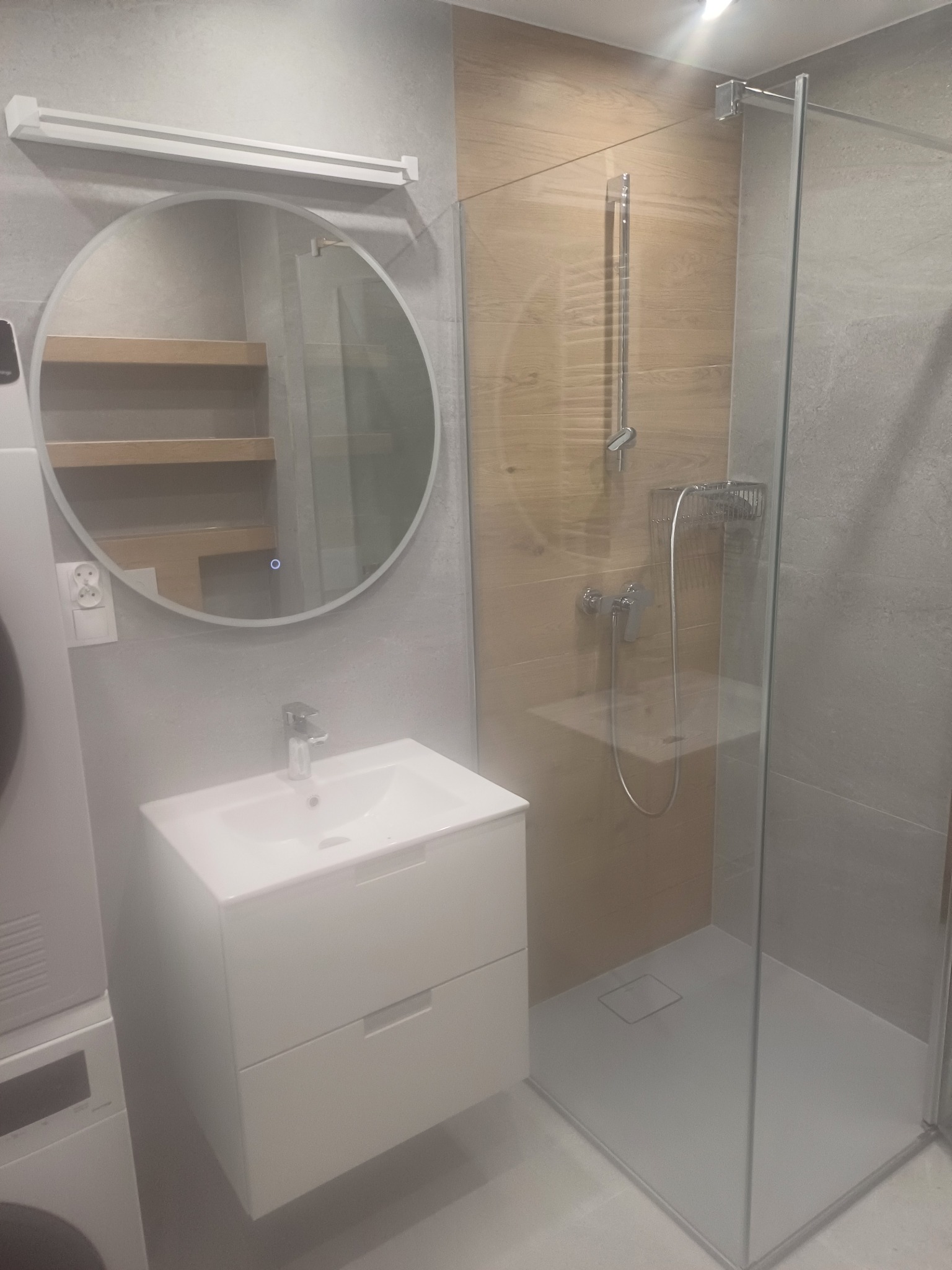 Nowoczesna łazienka z kabiną prysznicową typu walk-in, okrągłym lustrem z podświetleniem i białą szafką umywalkową z dwiema szufladami, widoczna pralka obok umywalki.