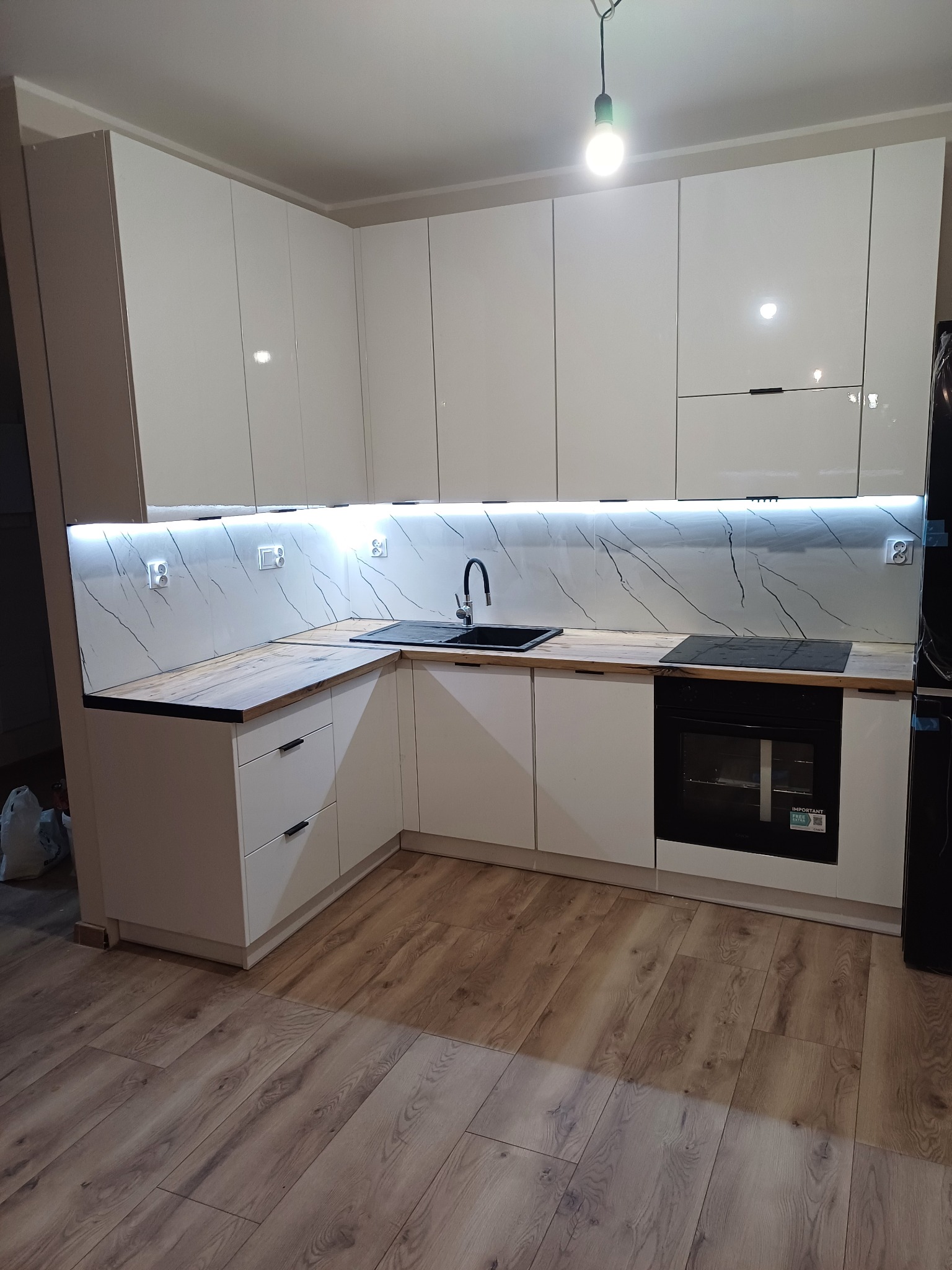 Nowoczesna kuchnia z białymi, lakierowanymi szafkami, drewnianym blatem i marmurowym panelem ściennym z podświetleniem LED, czarny zlew i płyta indukcyjna.