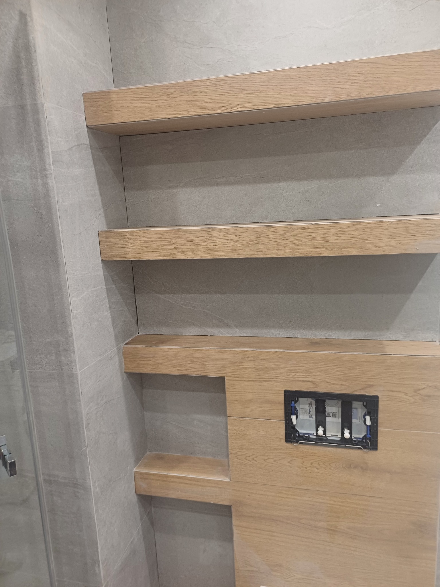 Półki z drewnianym wykończeniem w łazience wyłożonej szarymi płytkami, widoczne puszki elektryczne przed montażem osprzętu.