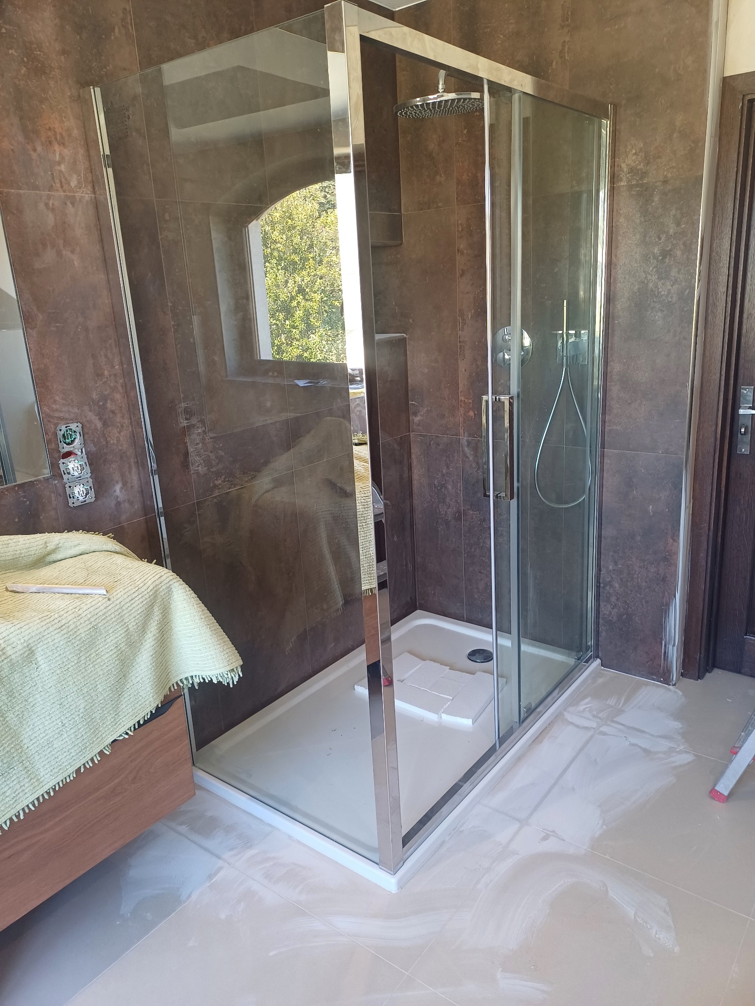 Kabina prysznicowa z przesuwnymi drzwiami, chromowanymi profilami i deszczownicą, zamontowana w łazience z brązowymi płytkami ściennymi. Na podłodze widoczne ślady po fugowaniu, a obok kabiny stoi...