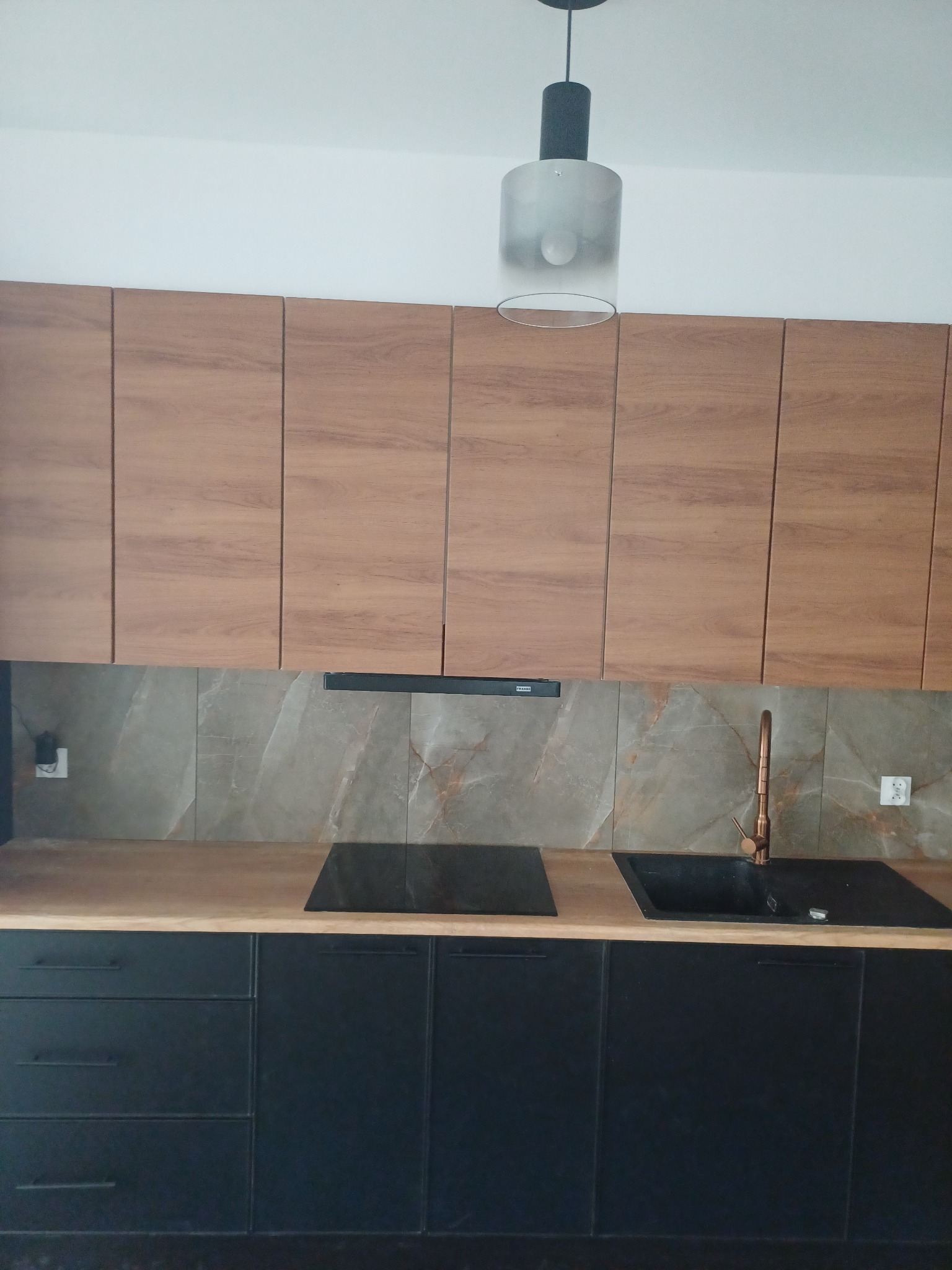 Nowoczesna kuchnia z drewnianymi górnymi szafkami, czarnymi dolnymi szafkami, marmurowymi płytkami na ścianie i miedzianą baterią.