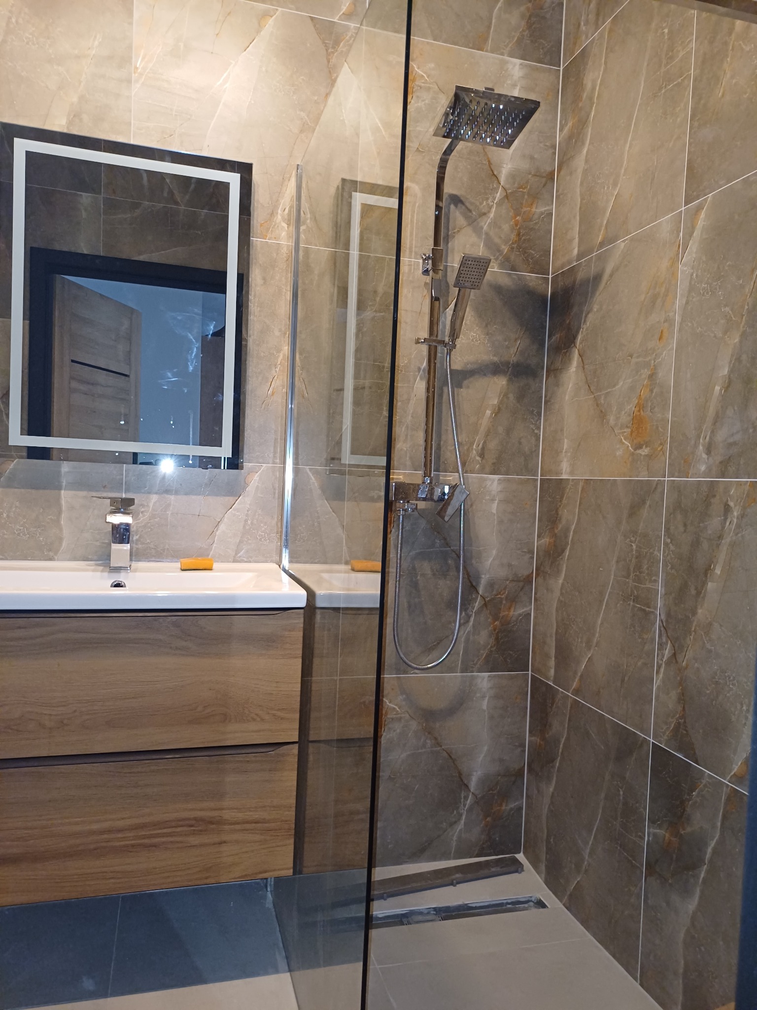 Nowoczesna łazienka z kabiną prysznicową typu walk-in, płytkami imitującymi marmur, umywalką z drewnianą szafką i lustrem z podświetleniem LED.