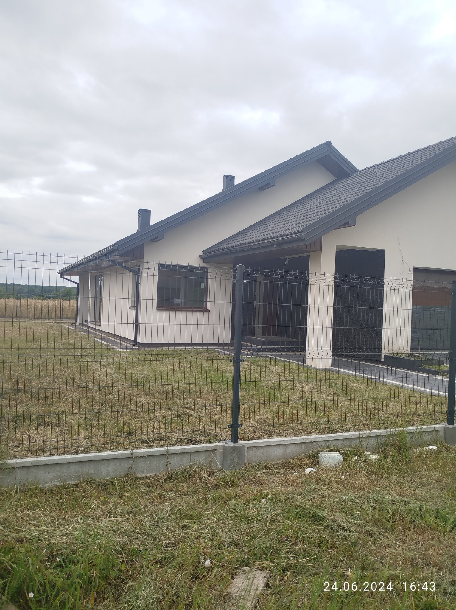 Jednorodzinny dom z jasną elewacją i ciemną dachówką, widoczny zza metalowego ogrodzenia panelowego na tle pochmurnego nieba i pola.