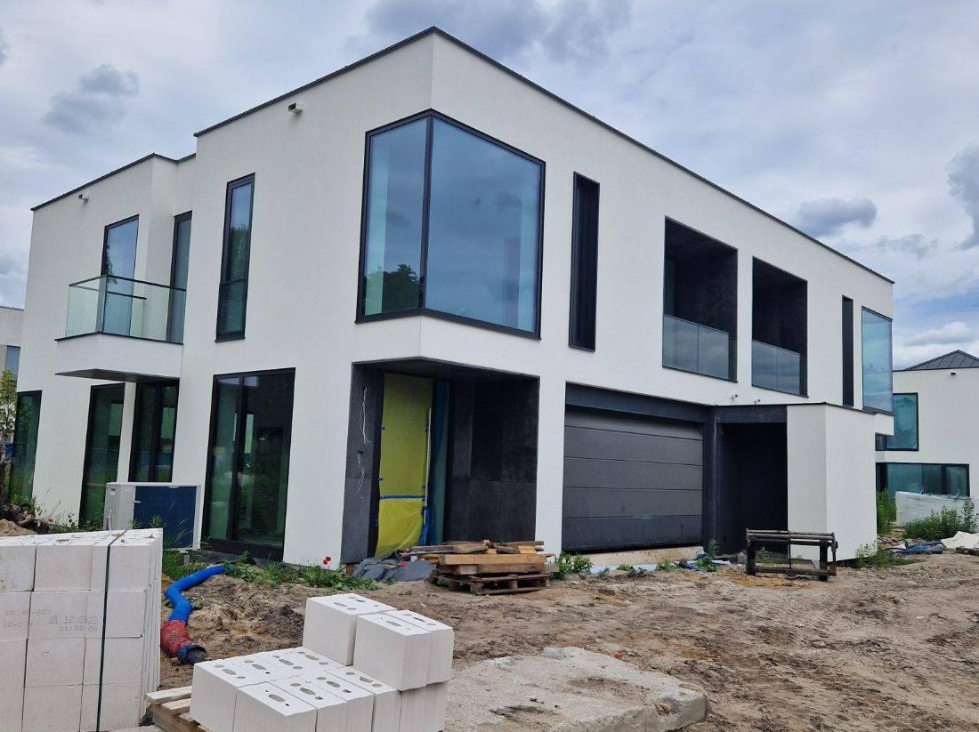 Nowoczesny, dwukondygnacyjny dom z białymi ścianami i dużymi, czarnymi oknami, w trakcie prac wykończeniowych; widoczne materiały budowlane i zabezpieczone wejście.