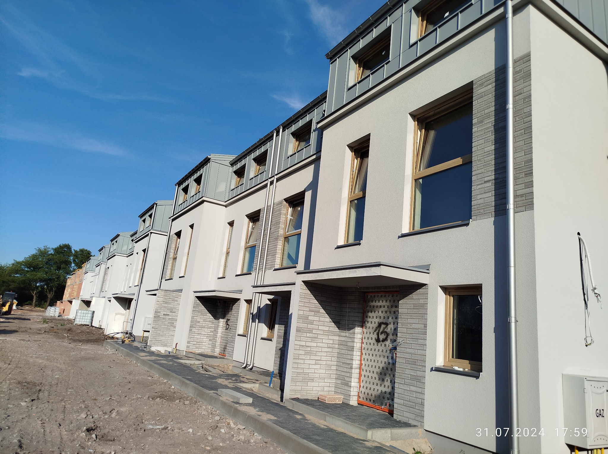 Szeregówka domów jednorodzinnych w trakcie wykończenia elewacji, widoczne tynki w odcieniach szarości, okna z drewnianą stolarką i metalowe obróbki blacharskie dachu, teren wokół budynków w trakcie...