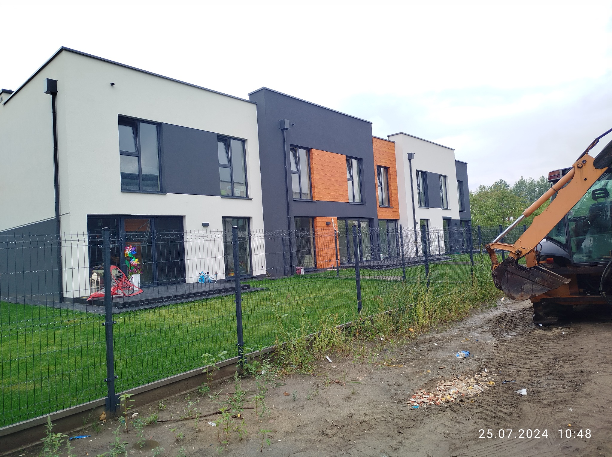 Nowoczesne szeregowe domy z elewacją w kolorach bieli, szarości i drewna, widoczne zza metalowego ogrodzenia, z koparką w prawym dolnym rogu.