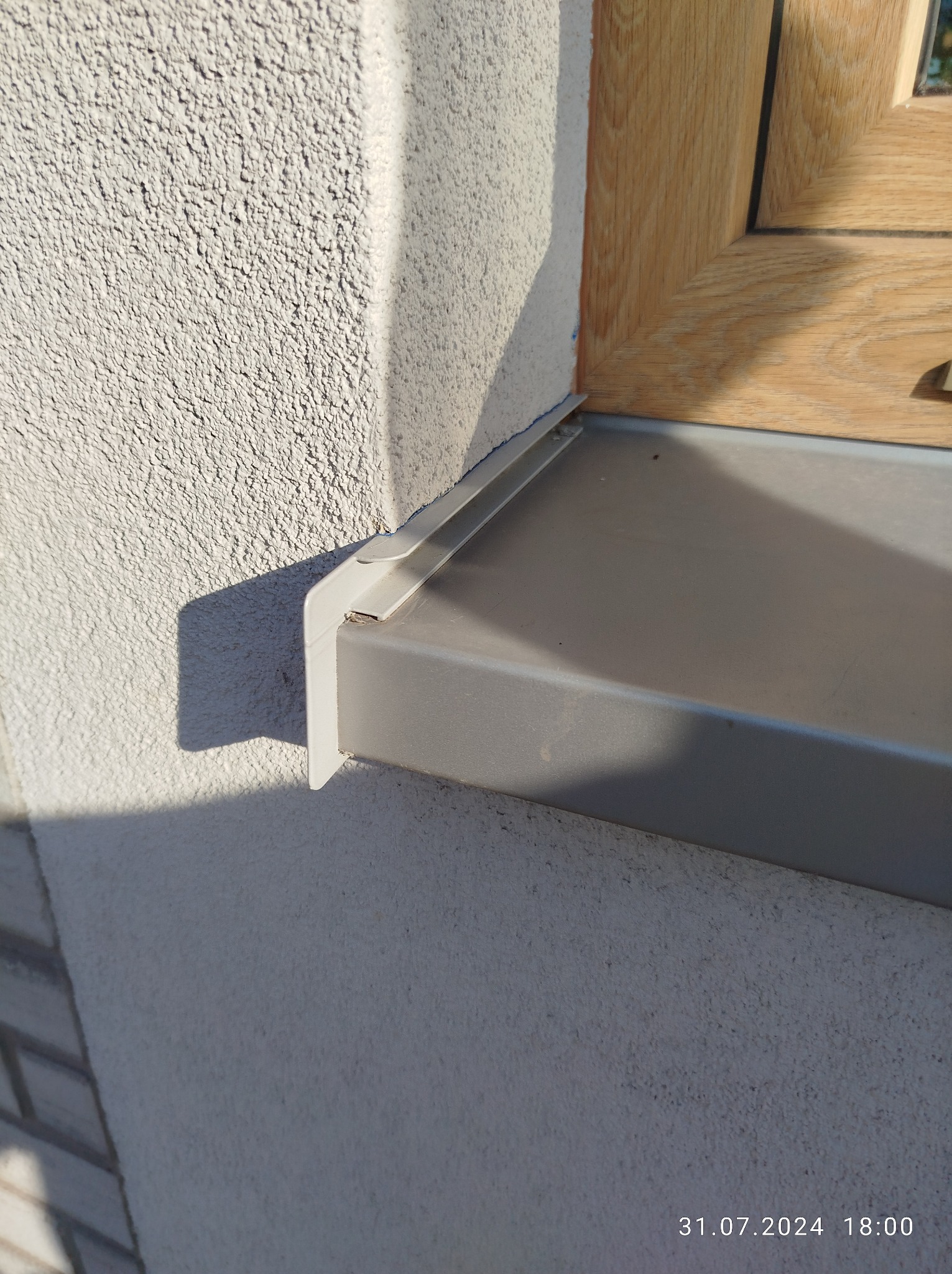 Detal elewacji z oknem: szary parapet okienny z metalowym wykończeniem, jasna ściana z fakturą baranka i drewniana rama okienna w ciepłym odcieniu.