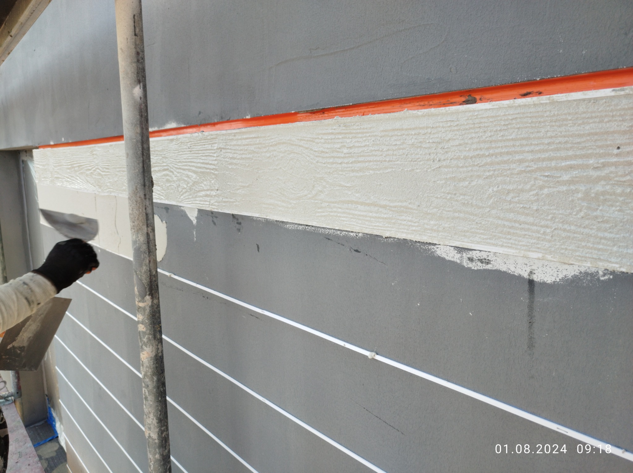 Nakładanie jasnej warstwy imitującej drewno na szarą elewację budynku, widoczny fragment rusztowania i pomarańczowa rurka pod sufitem.