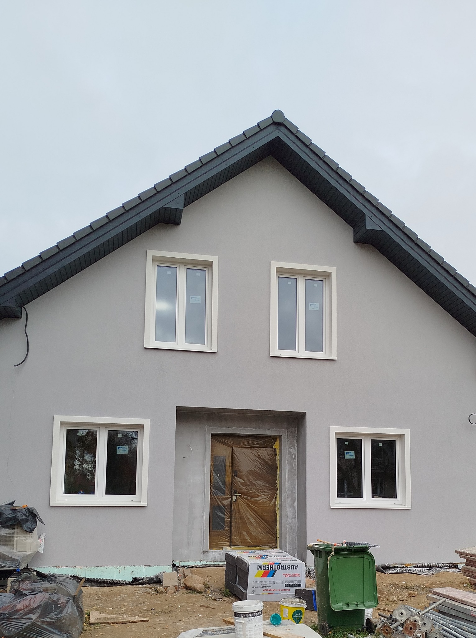 Dom jednorodzinny z szarą elewacją i białymi ramami okiennymi w trakcie prac wykończeniowych, widoczne materiały budowlane i zabezpieczone wejście.