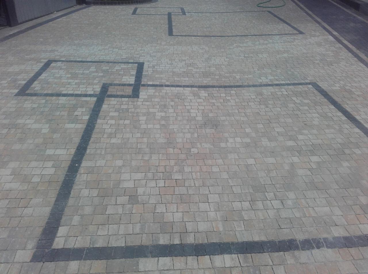 Ułożona kostka brukowa w odcieniach beżu i szarości, z ciemnymi obramowaniami tworzącymi geometryczne wzory kwadratów na powierzchni chodnika.