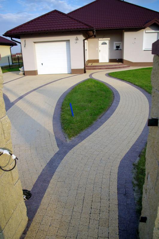 Wykonana z kostki brukowej, wijąca się ścieżka prowadząca do domu w Strzyżowie, z trawnikiem pośrodku i elementami małej architektury.