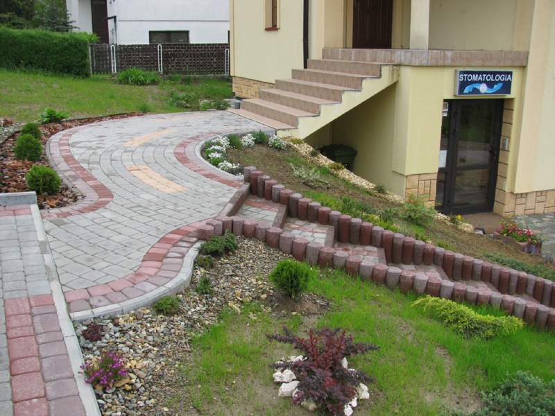 Kręty chodnik z kostki brukowej w odcieniach szarości i czerwieni, prowadzący do wejścia do gabinetu stomatologicznego; obrzeża chodnika wykończone dekoracyjnymi kamieniami i roślinami.
