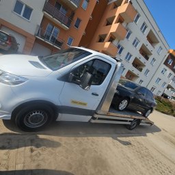 My dreams Justyna Kowalewska - Biała autolaweta z czarnym samochodem osobowym na platformie, zaparkowana na placu przed budynkiem mieszkalnym w słoneczny dzień. Widoczny napis 'Pomoc Drogowa'.