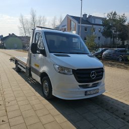 My dreams Justyna Kowalewska - Biały Mercedes-Benz Sprinter z platformą, zaparkowany na chodniku w słoneczny dzień. Widoczny napis 'Pomoc Drogowa' na boku pojazdu i budynki w tle.