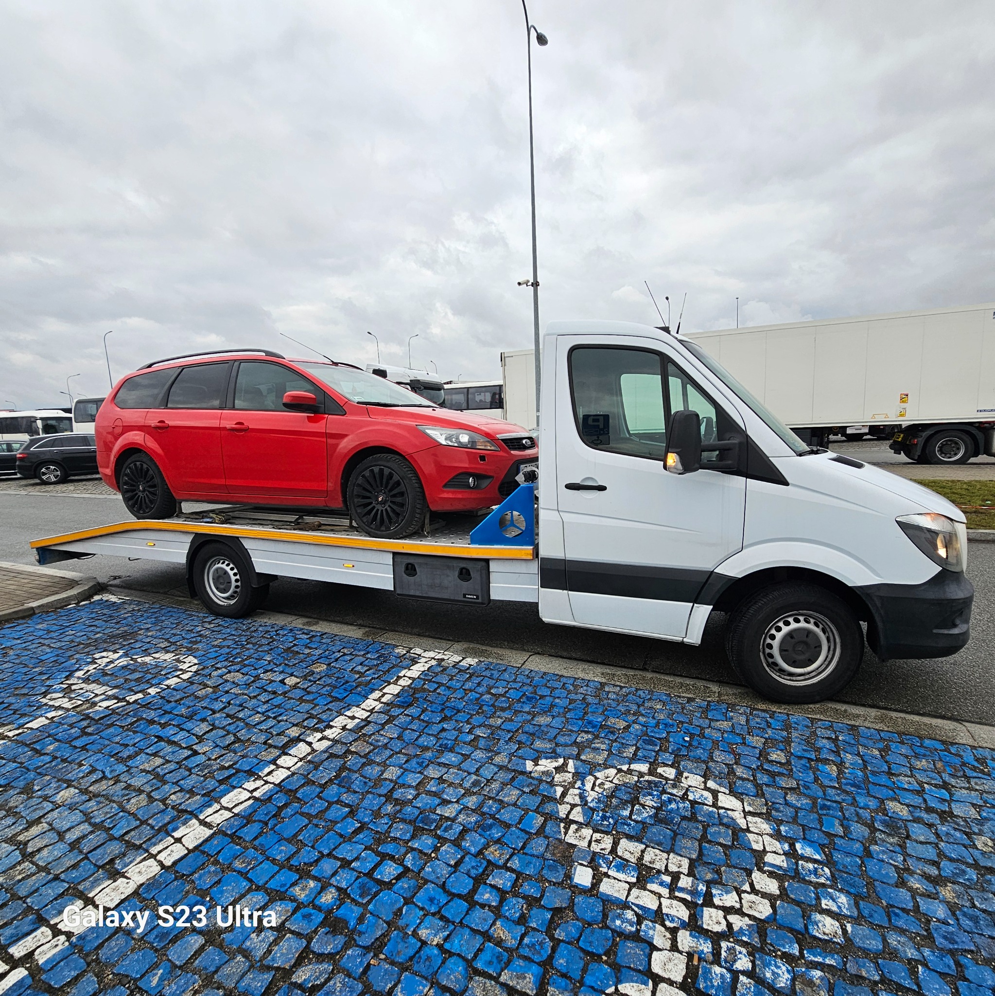 Czerwone kombi z czarnymi felgami transportowane na białym samochodzie z platformą na parkingu z kostki brukowej w niebiesko-białe wzory.