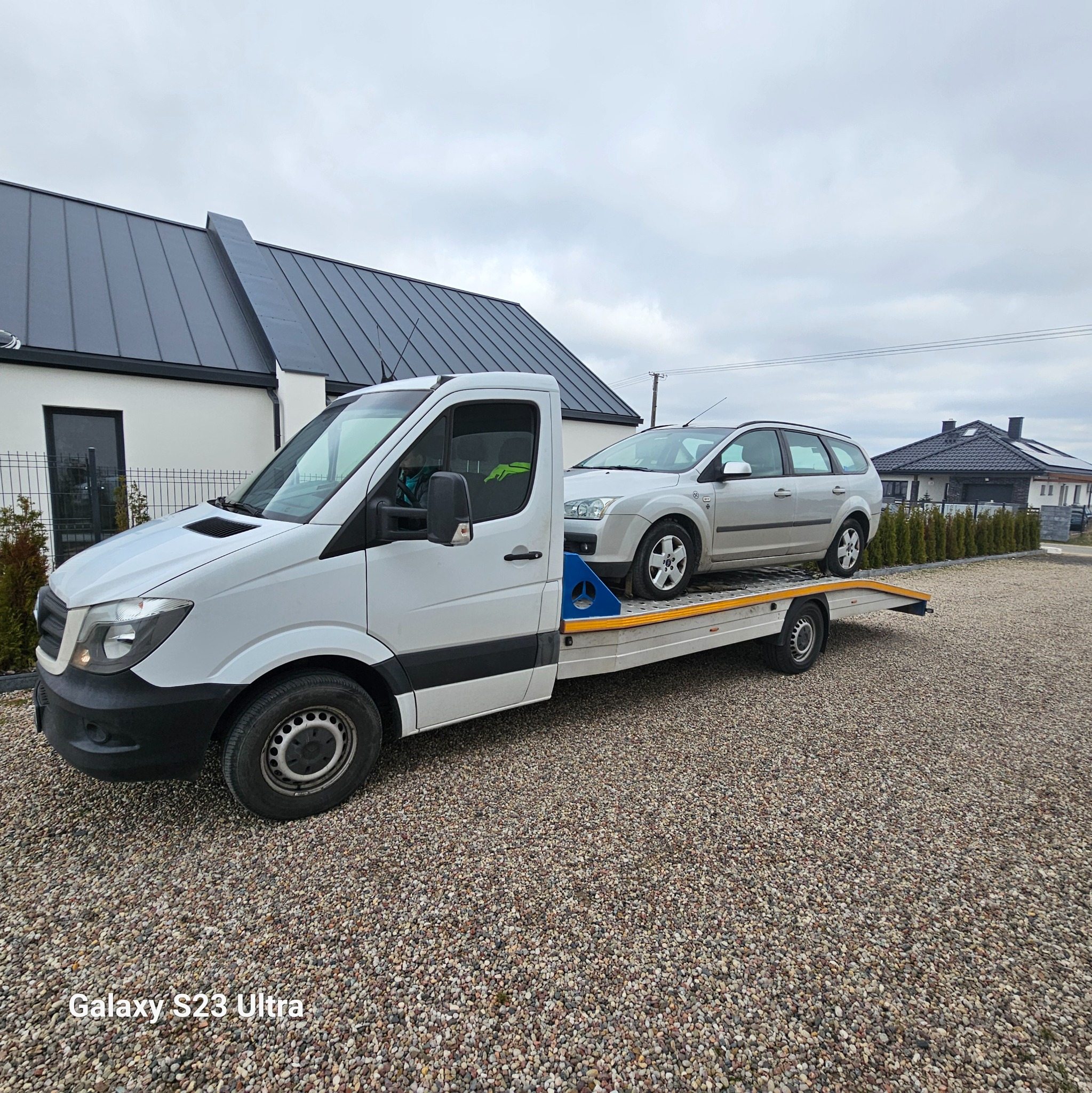 Biały samochód dostawczy z platformą transportową przewozi srebrne kombi na tle nowoczesnego domu z ciemnym dachem.