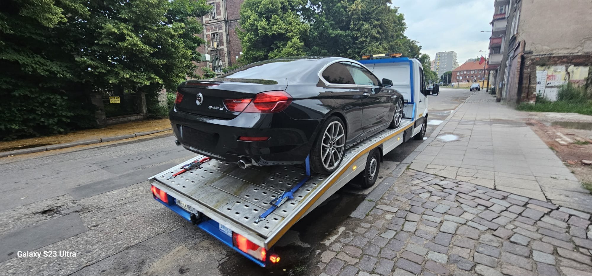 Czarny samochód BMW 640d zabezpieczony pasami transportowymi na platformie pojazdu pomocy drogowej, widok z tyłu, ulica miejska w tle.