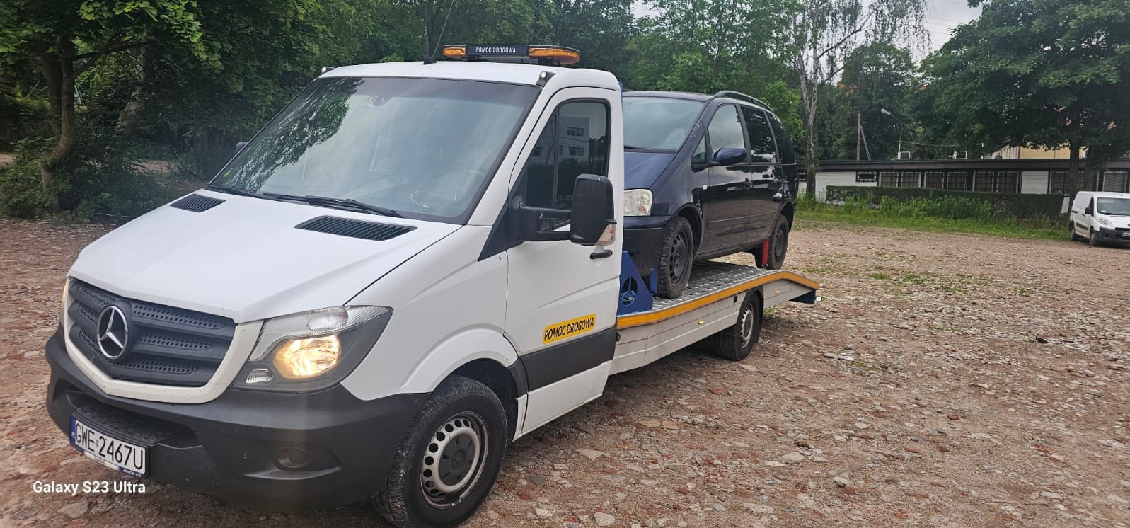 Biały samochód pomocy drogowej Mercedes Sprinter z niebieskim samochodem osobowym na platformie, zaparkowany na żwirowym placu, widoczny znak 'Pomoc Drogowa' na pojeździe.