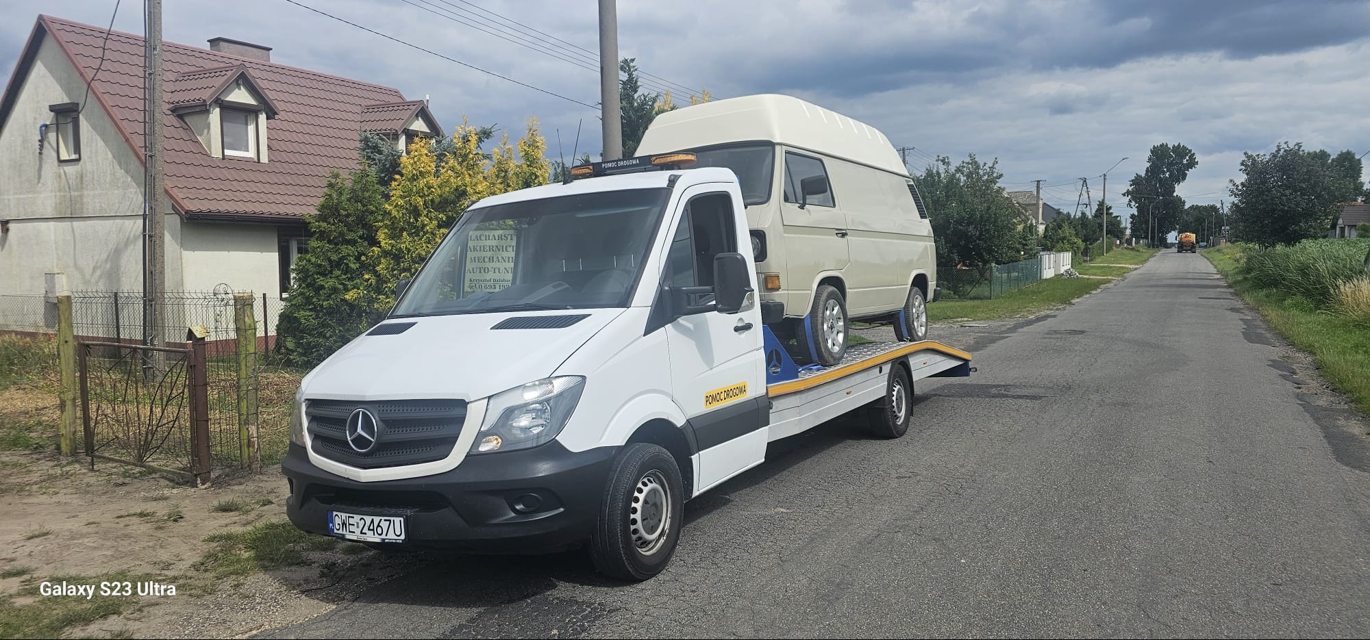 Biały Mercedes Sprinter z platformą transportową przewozi kremowy, podwyższony model Volkswagena T3 na wiejskiej drodze, z domami w tle i tablicą informacyjną z nazwą firmy na kabinie.