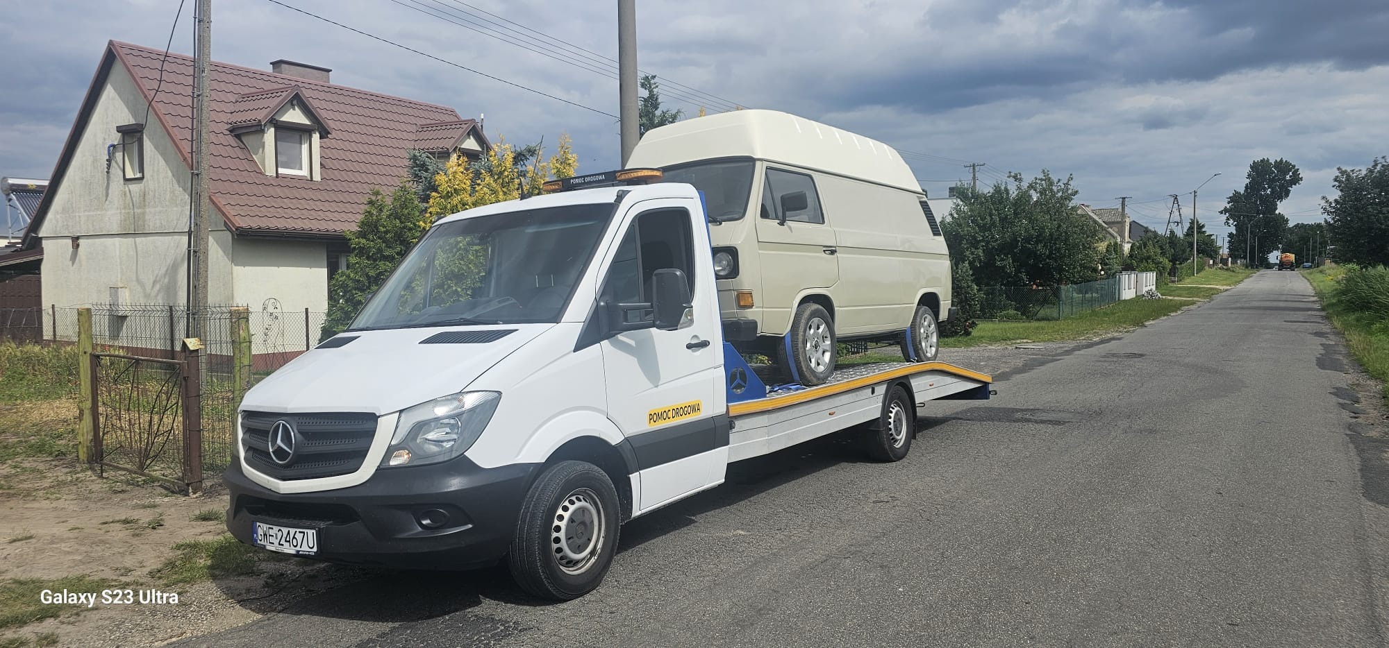 Biała ciężarówka pomocy drogowej Mercedes-Benz z żółtym napisem 'POMOC DROGOWA' przewozi beżowego Volkswagena Transportera T3 z wysokim dachem na platformie, na tle wiejskiego krajobrazu z domami...