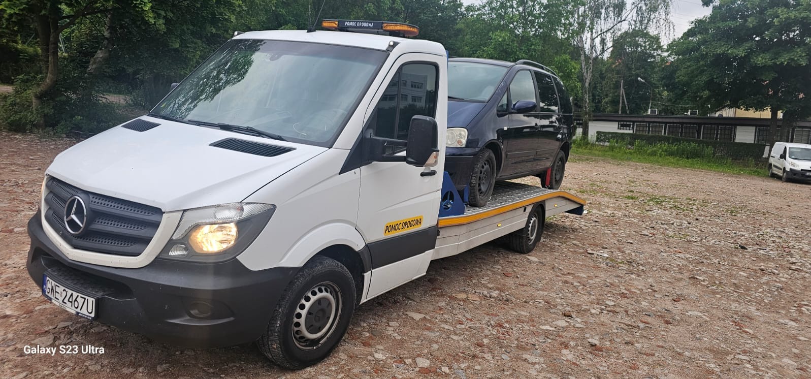 Biała laweta Mercedes z niebieskim samochodem osobowym na platformie, widoczny napis 'Pomoc Drogowa' i pomarańczowe światło ostrzegawcze na dachu, na szutrowym placu.