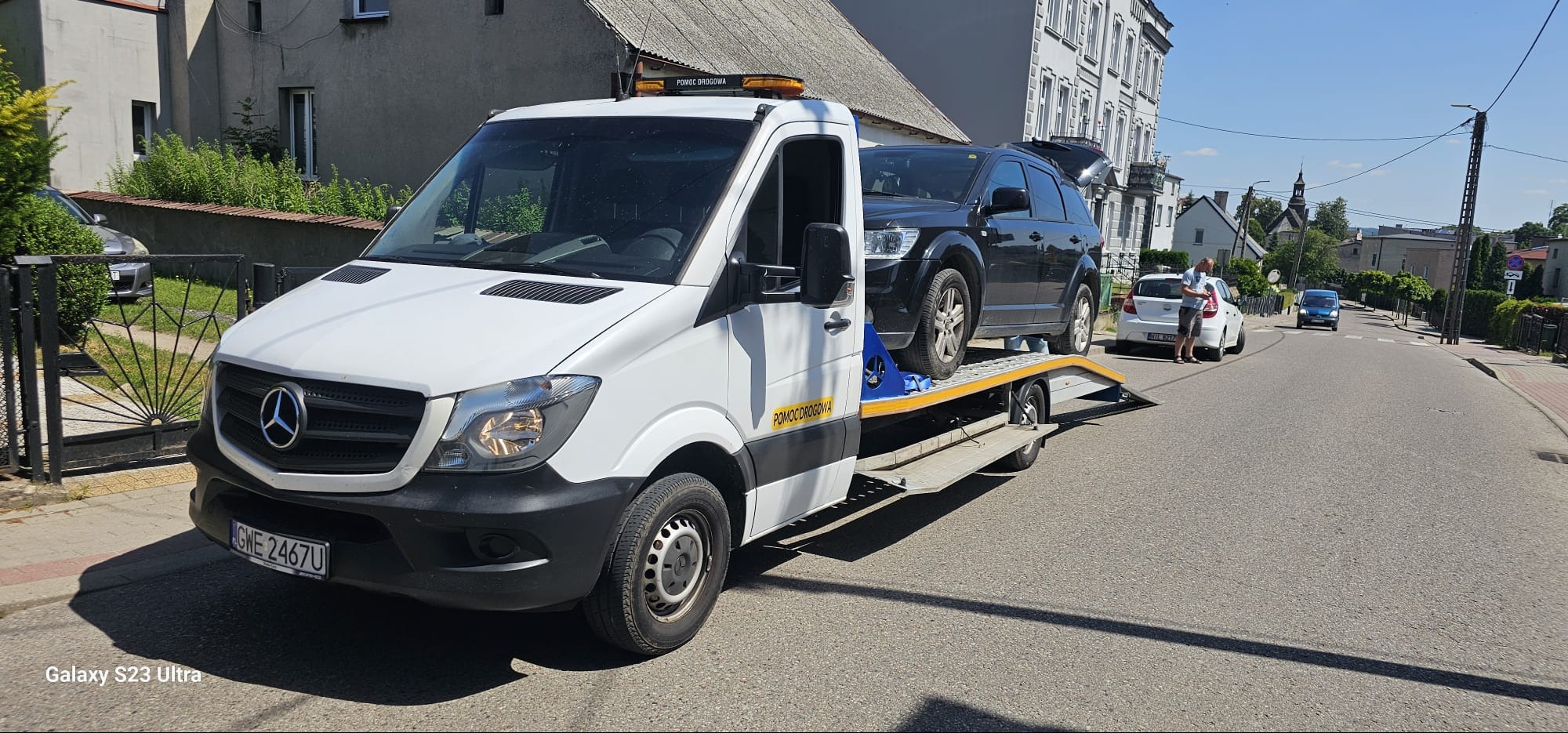 Biały Mercedes Sprinter z napisem 'POMOC DROGOWA' przewozi czarnego SUV-a na platformie w zabudowie w słoneczny dzień na ulicy.