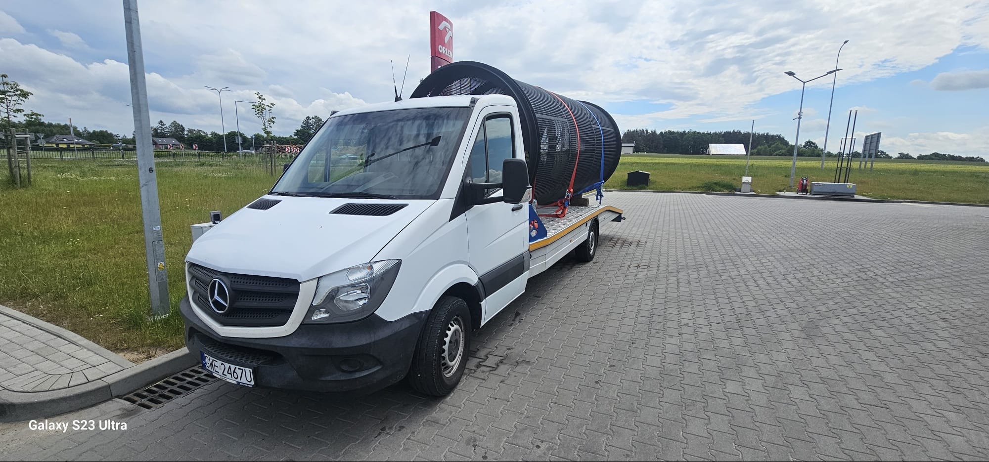 Biały Mercedes Sprinter z platformą transportową przewozi duży, czarny, cylindryczny element zabezpieczony pasami, na tle stacji paliw Orlen i krajobrazu.