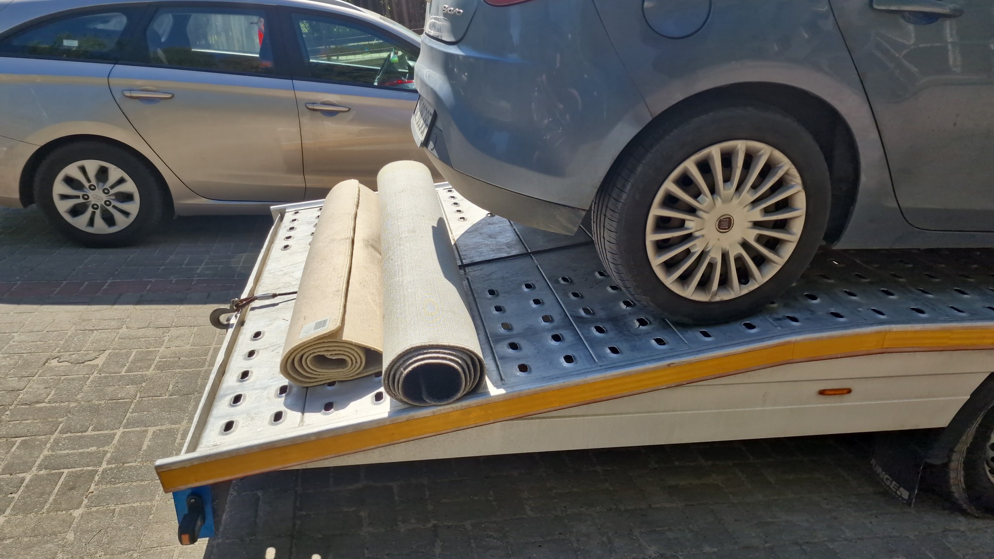 Samochód osobowy marki Fiat częściowo umieszczony na platformie transportowej z dwoma zwiniętymi dywanami, widoczne koło z logo producenta.