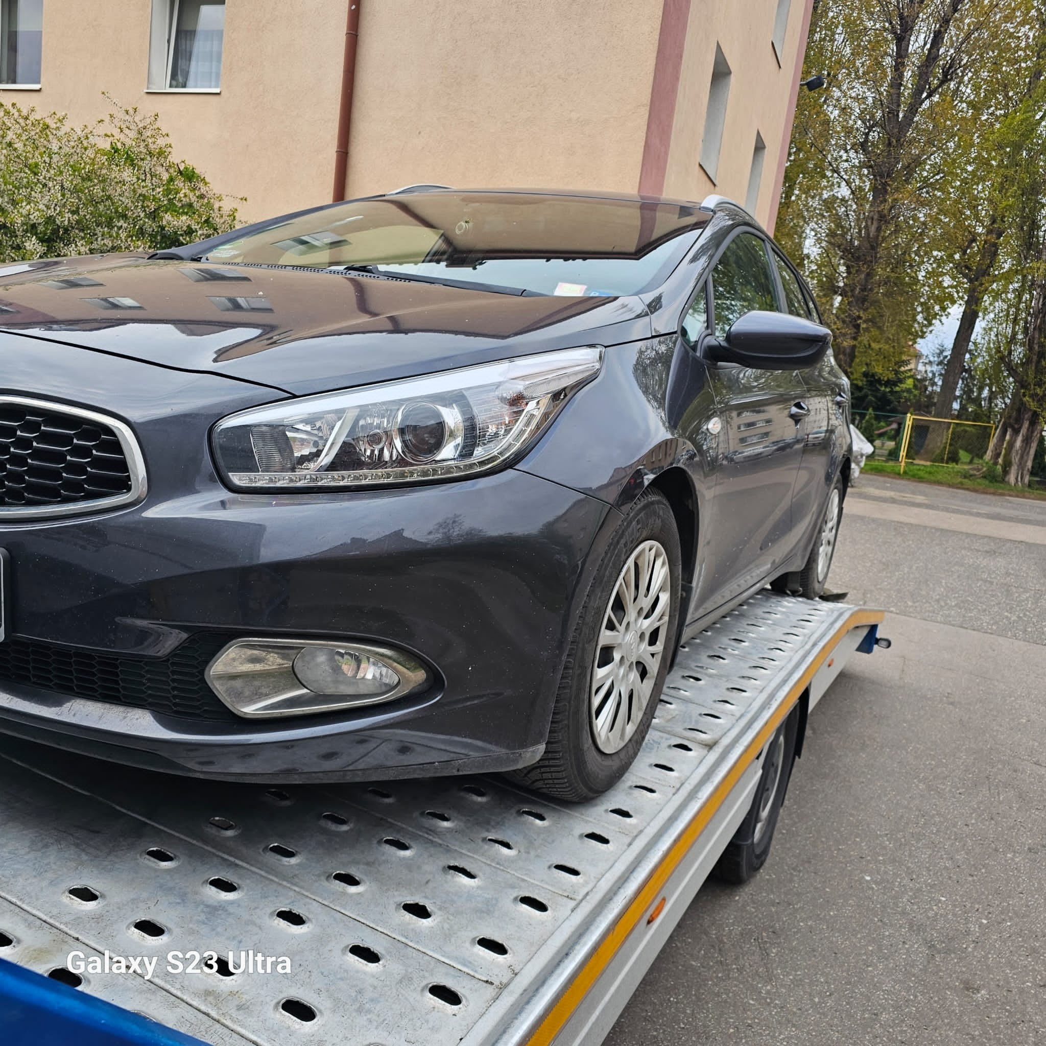 Szary samochód osobowy marki Kia załadowany na platformę pojazdu transportowego, widok z przodu, w tle budynek mieszkalny i drzewa.