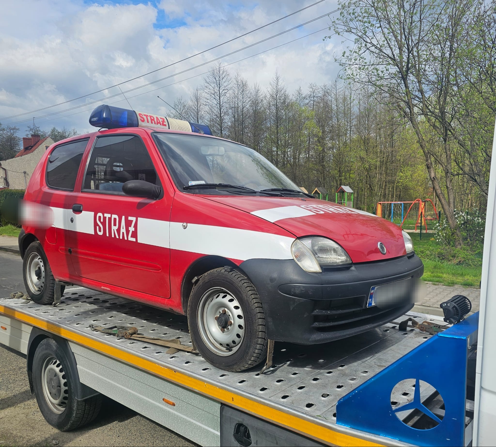 Czerwony samochód straży pożarnej z białym pasem i napisem 'STRAŻ' przewożony na platformie transportowej w słoneczny dzień, w tle widoczny plac zabaw i drzewa.