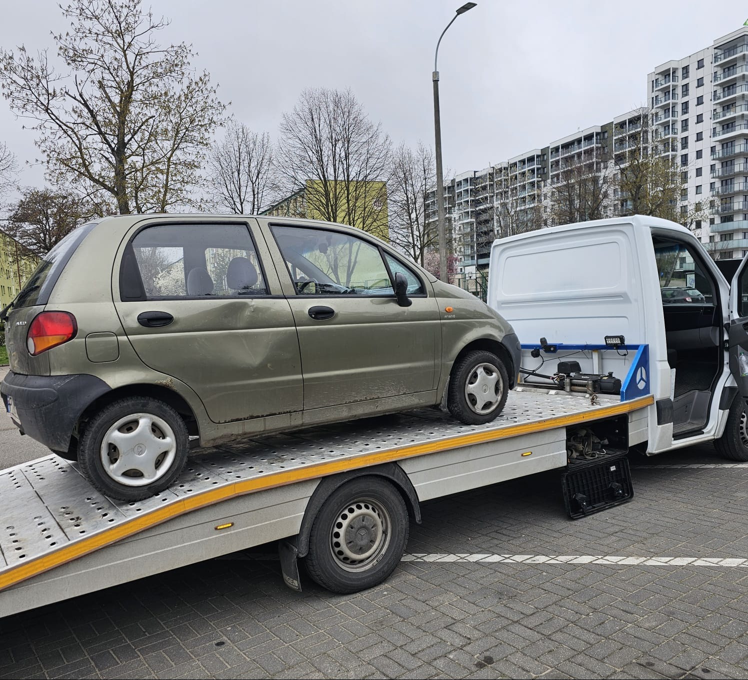 Uszkodzony, oliwkowy samochód osobowy Daewoo Matiz transportowany na platformie białego pojazdu pomocy drogowej na tle osiedla mieszkaniowego w pochmurny dzień.