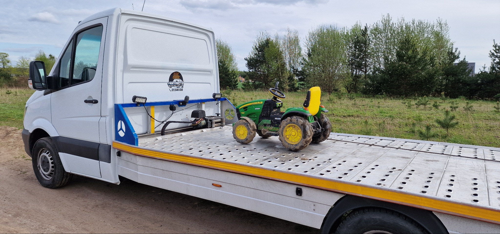 Biała laweta z niebieskimi elementami zabezpieczającymi transportuje zabrudzony, zielono-żółty, dziecięcy traktor John Deere na tle łąki i drzew.