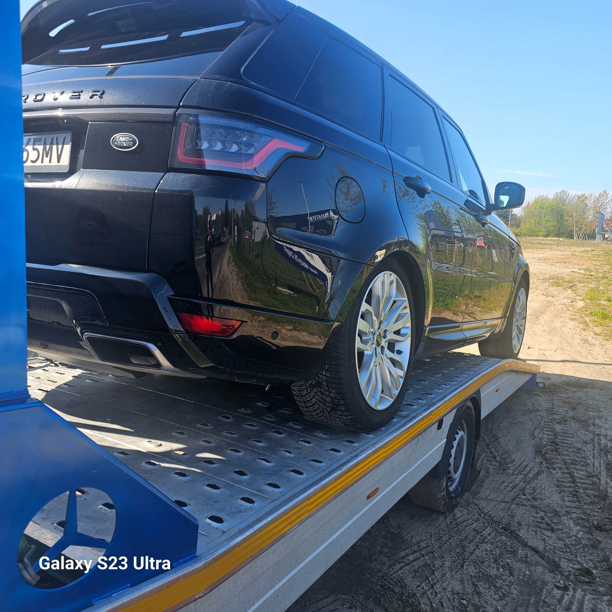 Czarny Range Rover Sport na platformie pomocy drogowej, widok z tyłu, srebrne felgi, tablica rejestracyjna, słoneczny dzień, teren poza miastem.