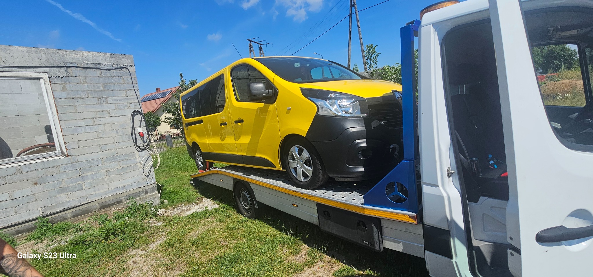 Żółty Renault Trafic na platformie białej lawety, widok z boku na tle budynku i zieleni. Słoneczny dzień, błękitne niebo.