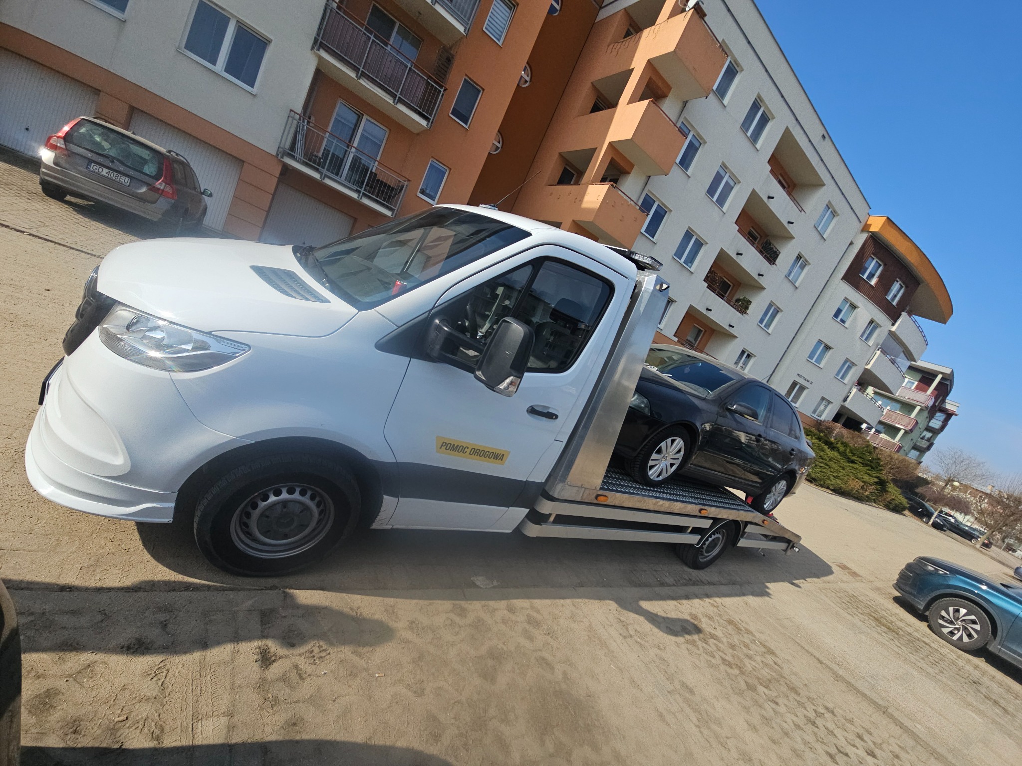 Biała autolaweta z czarnym samochodem osobowym na platformie, zaparkowana na placu przed budynkiem mieszkalnym w słoneczny dzień. Widoczny napis 'Pomoc Drogowa'.